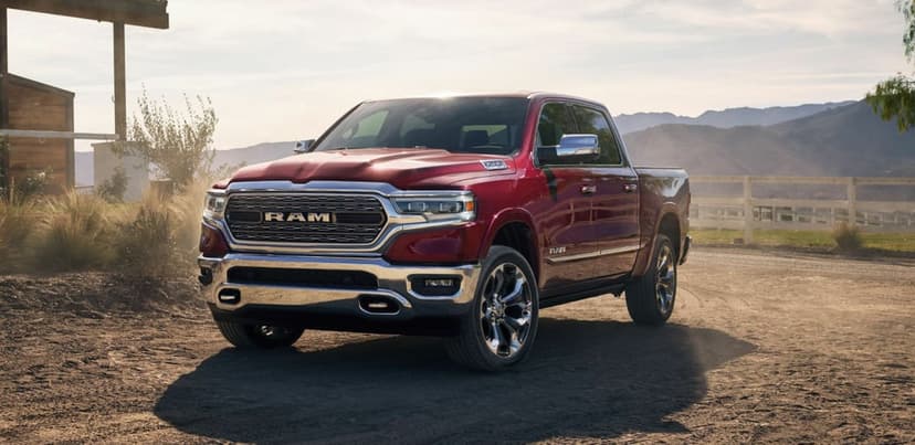 Red Ram 1500 pickup truck on a dusty road with chrome accents, wooden ranch buildings, and mountain scenery.