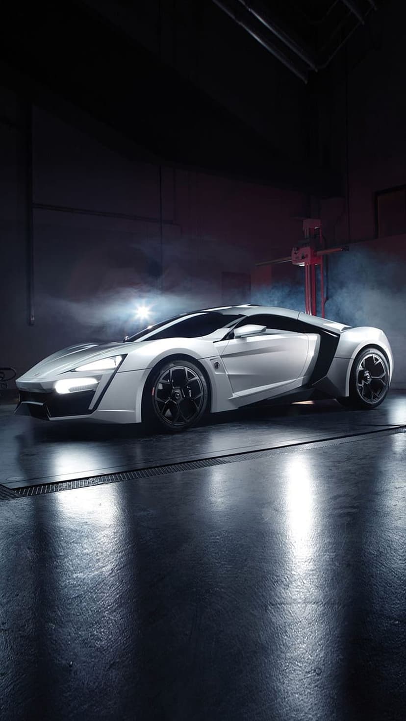 White hypercar with glowing headlights stands in a smoky studio on a reflective floor with black custom wheels.