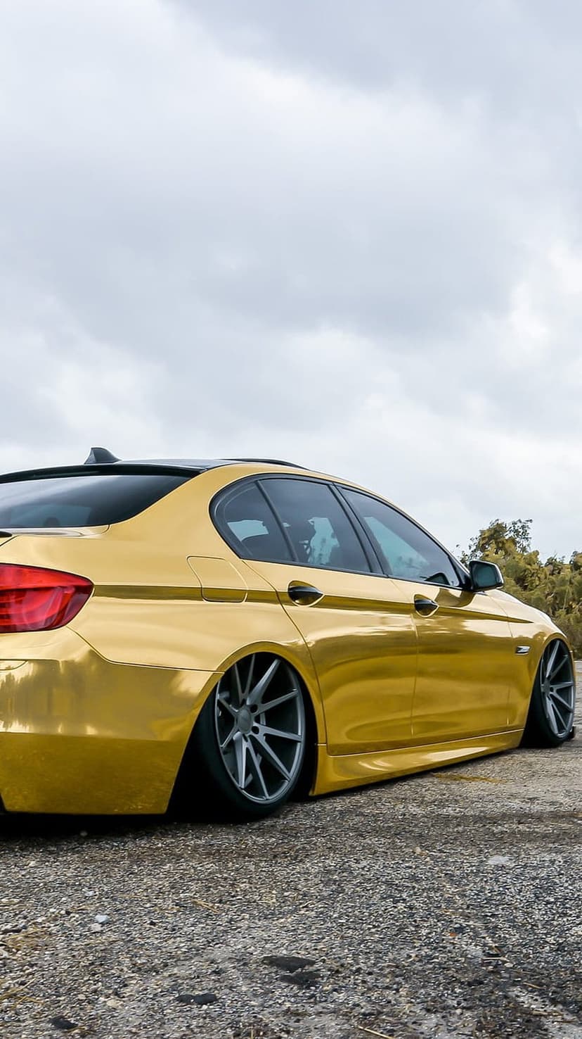 Gold BMW sedan with lowered suspension and grey multi-spoke rims parked on gravel under a cloudy sky.
