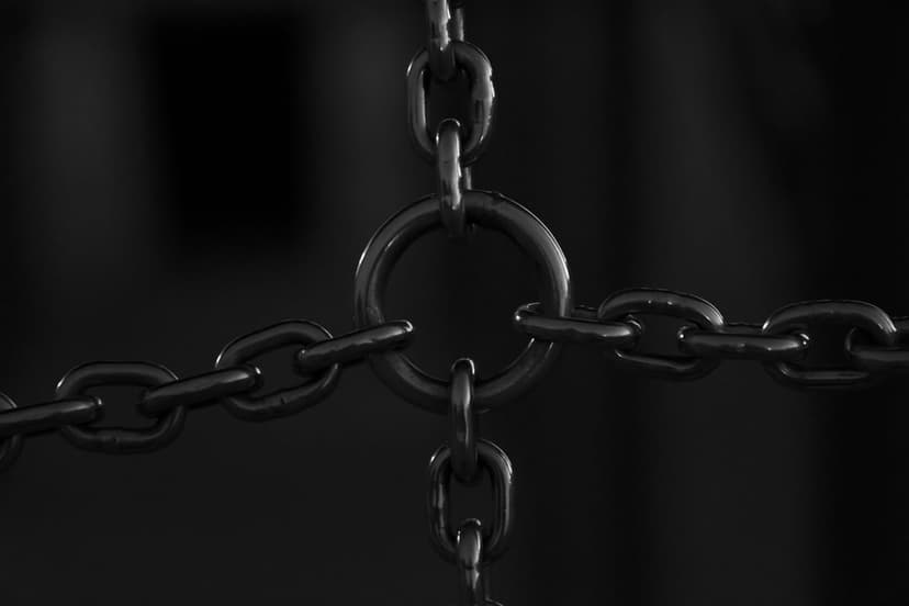 Interlocking metal chains hang from a central ring against a dark, out-of-focus industrial background in grayscale.