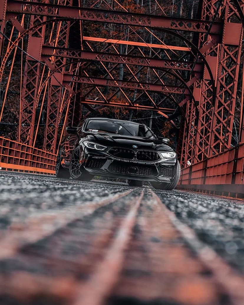 Black BMW M8 sports car parked on a red steel truss bridge with a low-angle view and weathered road texture.