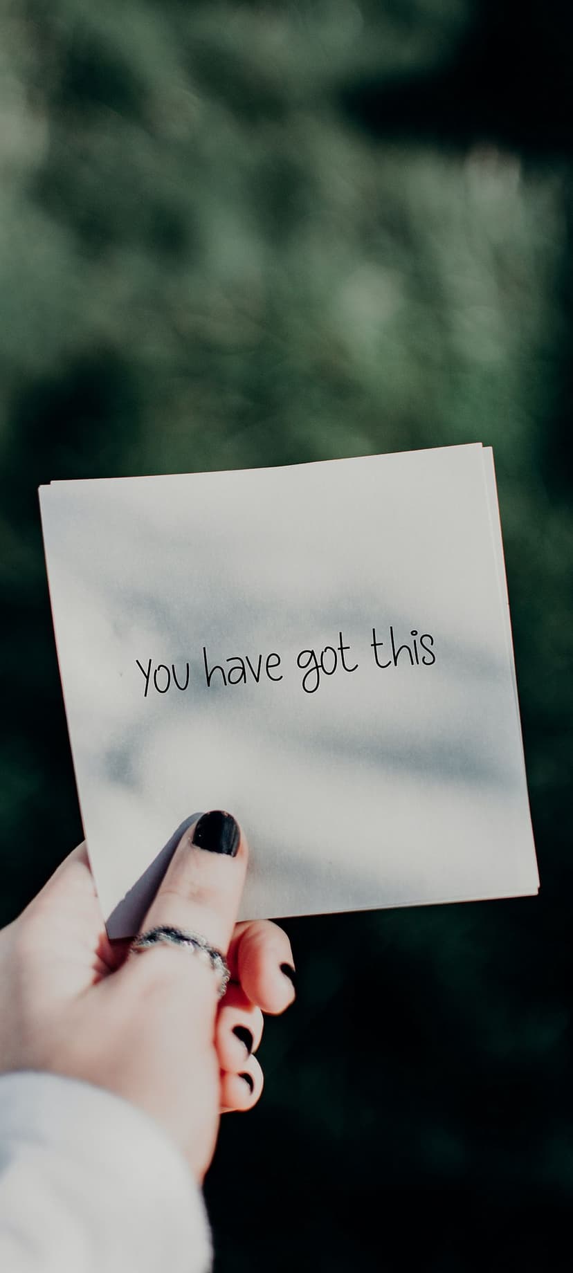 Hand with black nail polish holds a You Have Got This note over blurred dark green leaves in a forest setting.