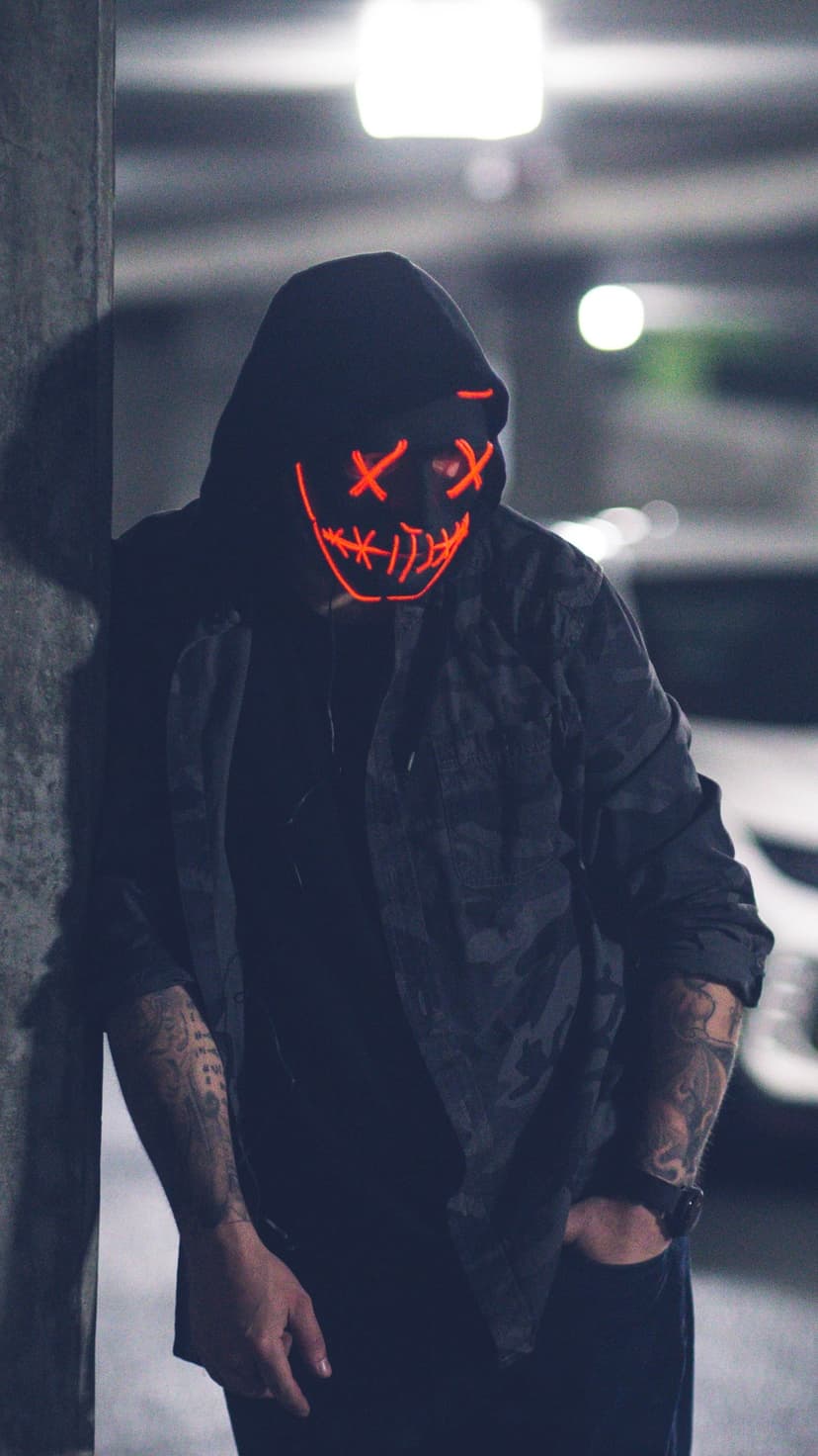 Person in a red glowing X-eye mask and dark hoodie leans against a concrete pillar in a dimly lit garage.