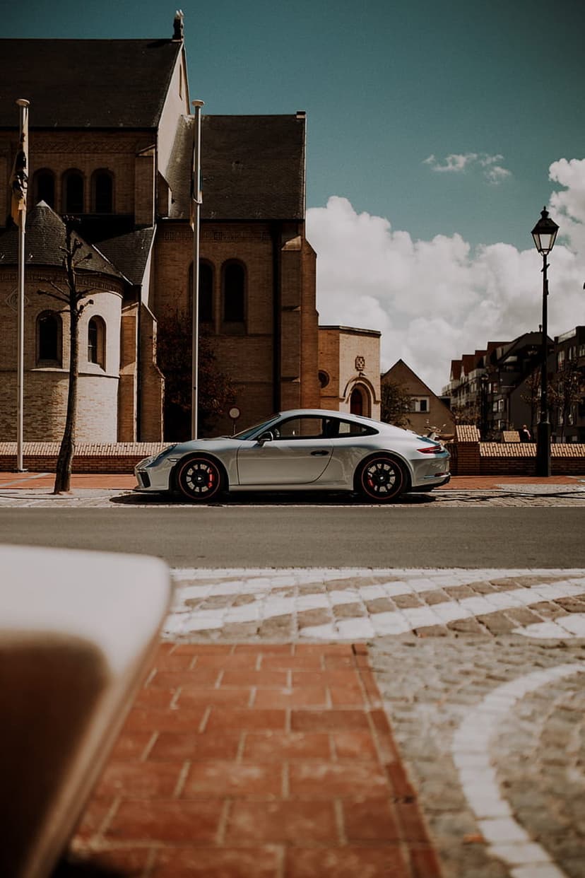 Silver Porsche 911 parked on a cobblestone street in front of a tan brick church under a bright blue sunny sky.