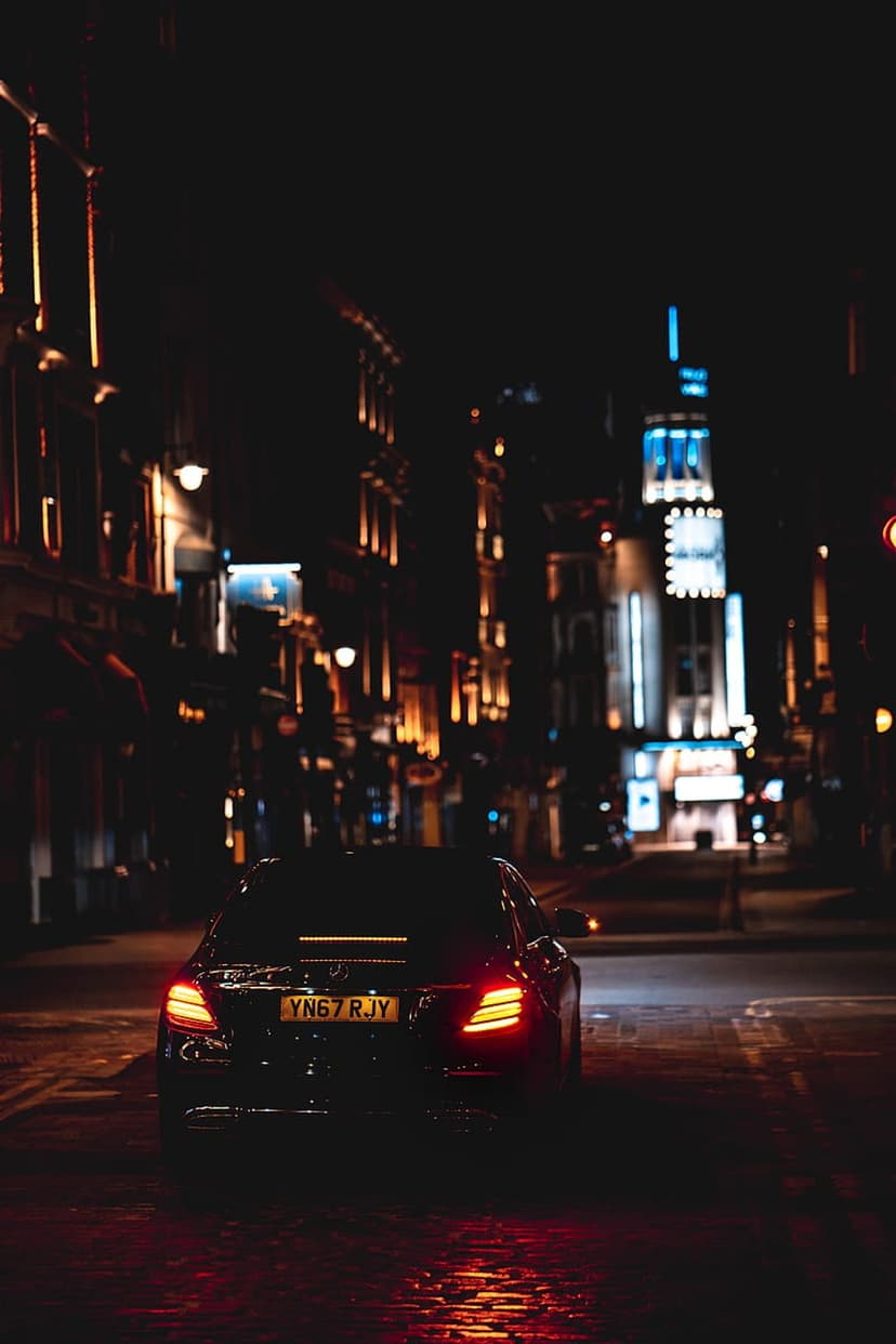 Black car with glowing red taillights parked on a wet city street between illuminated high-rise buildings at night.