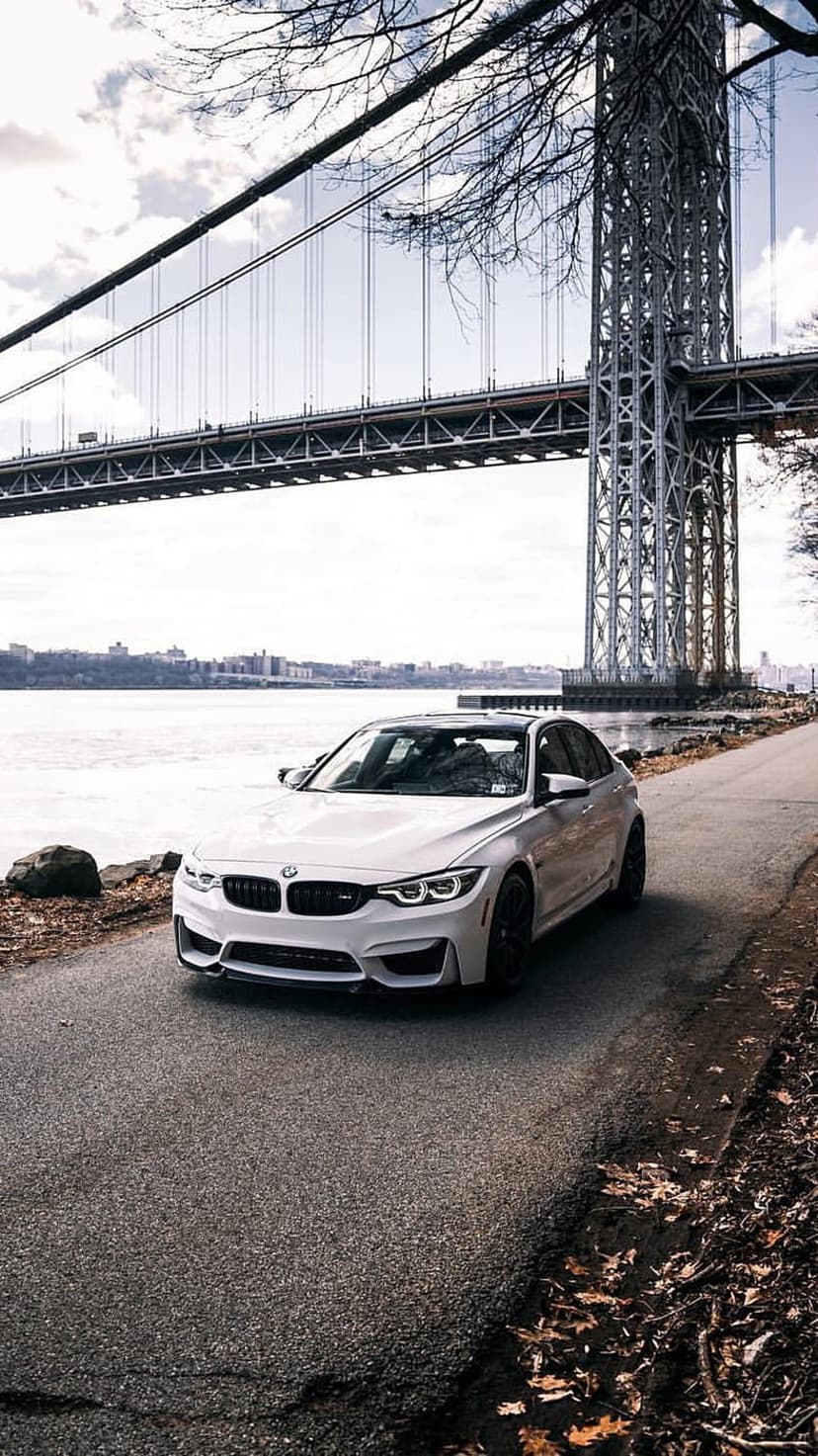 White BMW M3 sedan parked on an asphalt road beside a river with a large suspension bridge in the background.