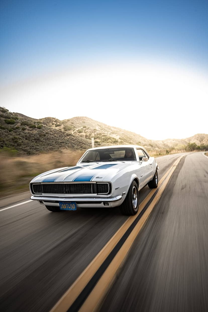 White classic Camaro with blue racing stripes speeds down a winding highway during a warm golden sunset.