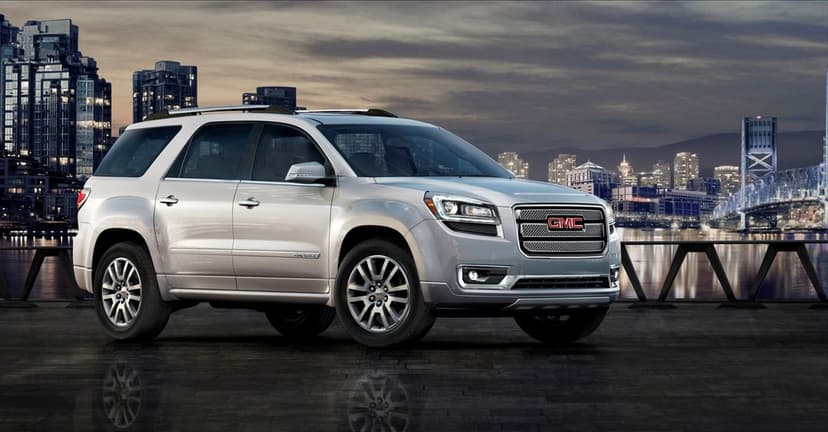 Silver GMC Acadia Denali SUV parked on a reflective surface with a lit city skyline and bridge at dusk.