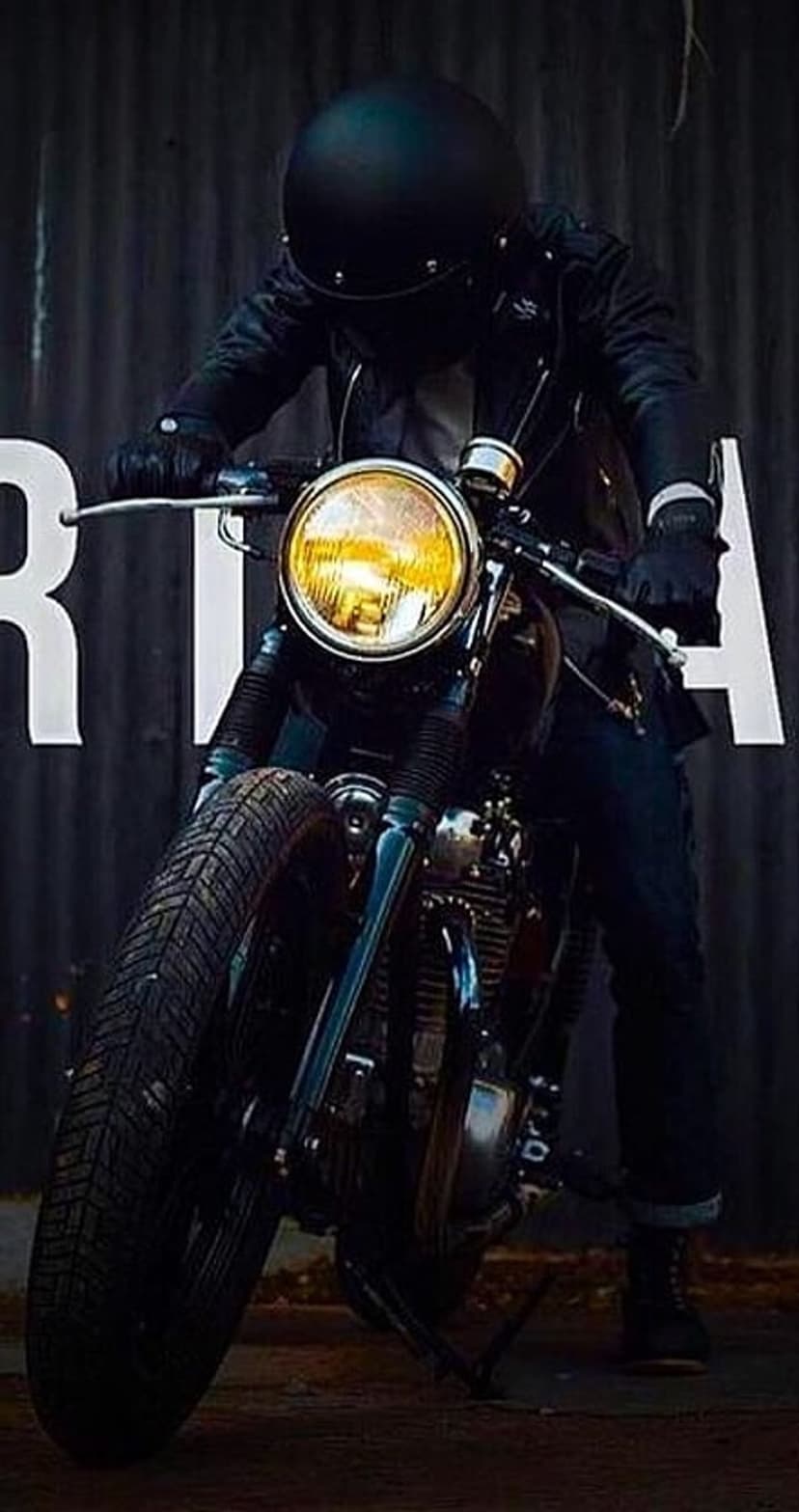 Biker in a black helmet on a cafe racer with a glowing yellow headlight against a dark corrugated metal wall.