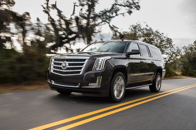 Dark Cadillac Escalade ESV driving on an asphalt road with motion blur and mossy trees in the background.
