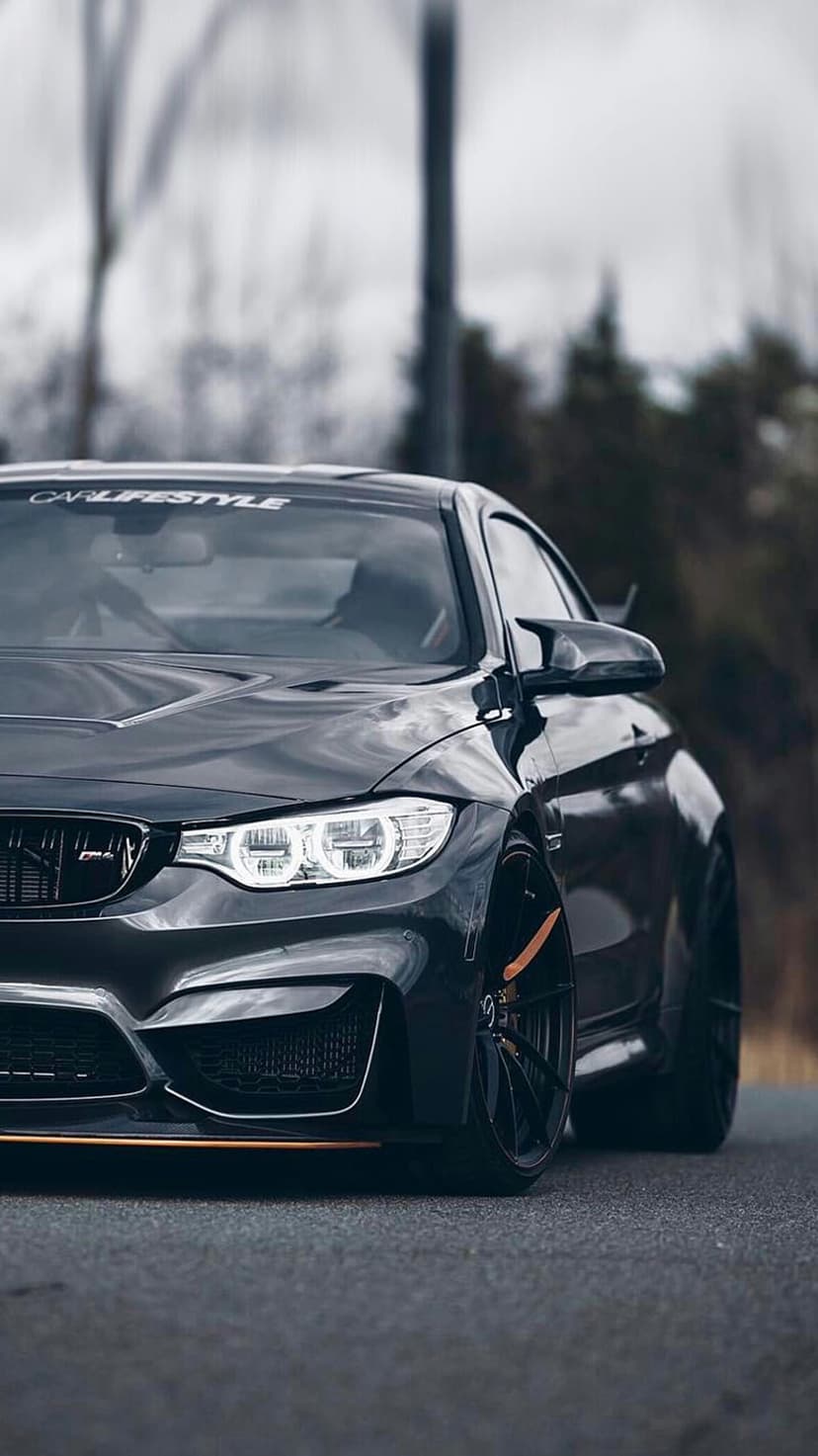 Black BMW M4 coupe with LED headlights and orange brake calipers parked on a road with a blurred forest backdrop.