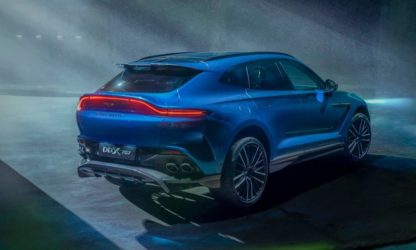 Rear view of a blue Aston Martin DBX 707 SUV in a dark studio with cinematic light beams and red taillights.