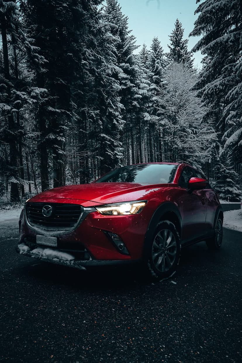 Red Mazda CX-3 SUV parked on a wet road amidst snow-covered pine trees with its headlights shining brightly.