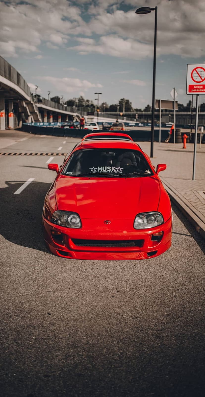 Red Toyota Supra sports car parked on a textured asphalt race track under bright sunlight with a blurred background.