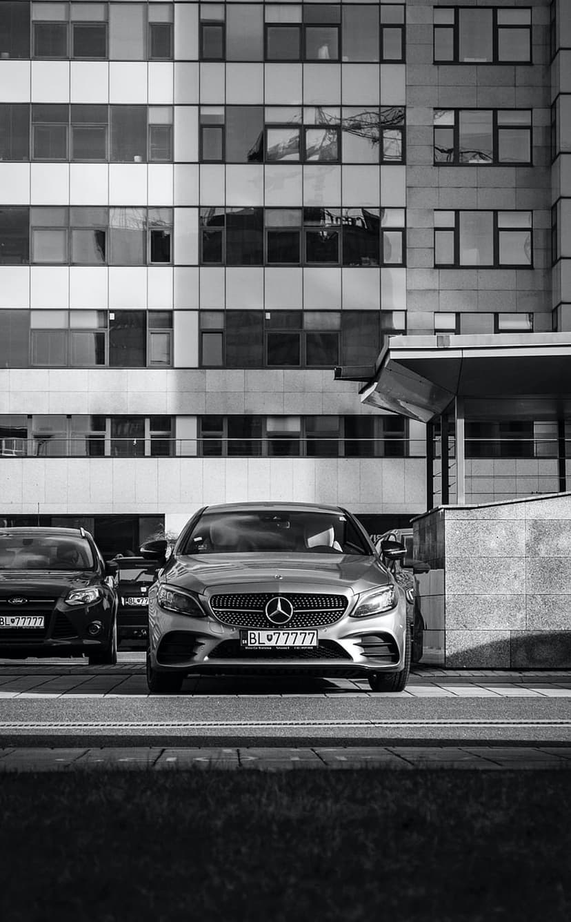 Mercedes-Benz and Ford sedans parked on a street outside a glass office building in a black and white urban scene.
