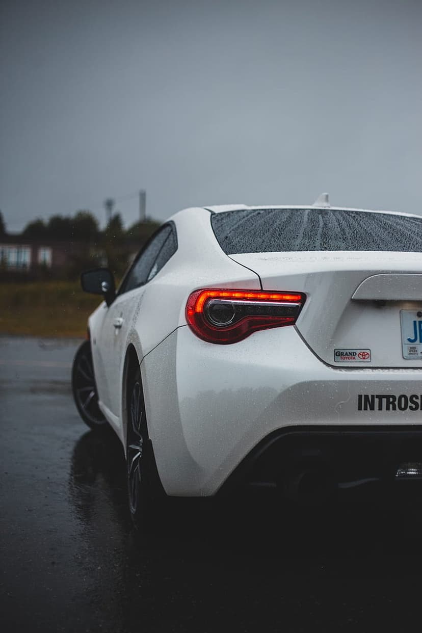 White Toyota GT86 rear tail lights glowing on wet asphalt during a gray rainy day for a mobile background.