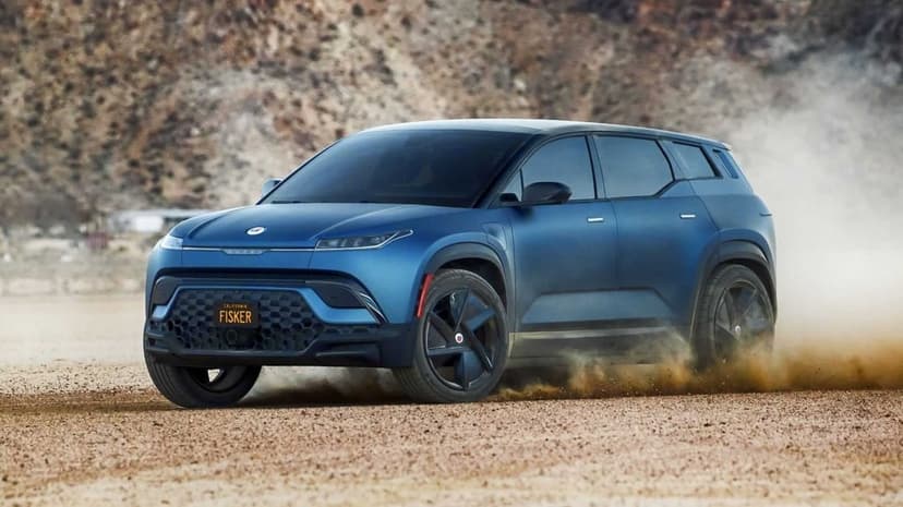 Blue Fisker Ocean SUV driving on a dirt road kicking up a cloud of dust against a rocky desert background.