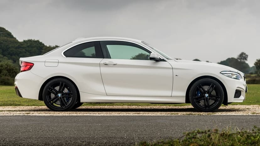 White BMW coupe side profile with black wheels and blue brake calipers on a road under a cloudy sky.
