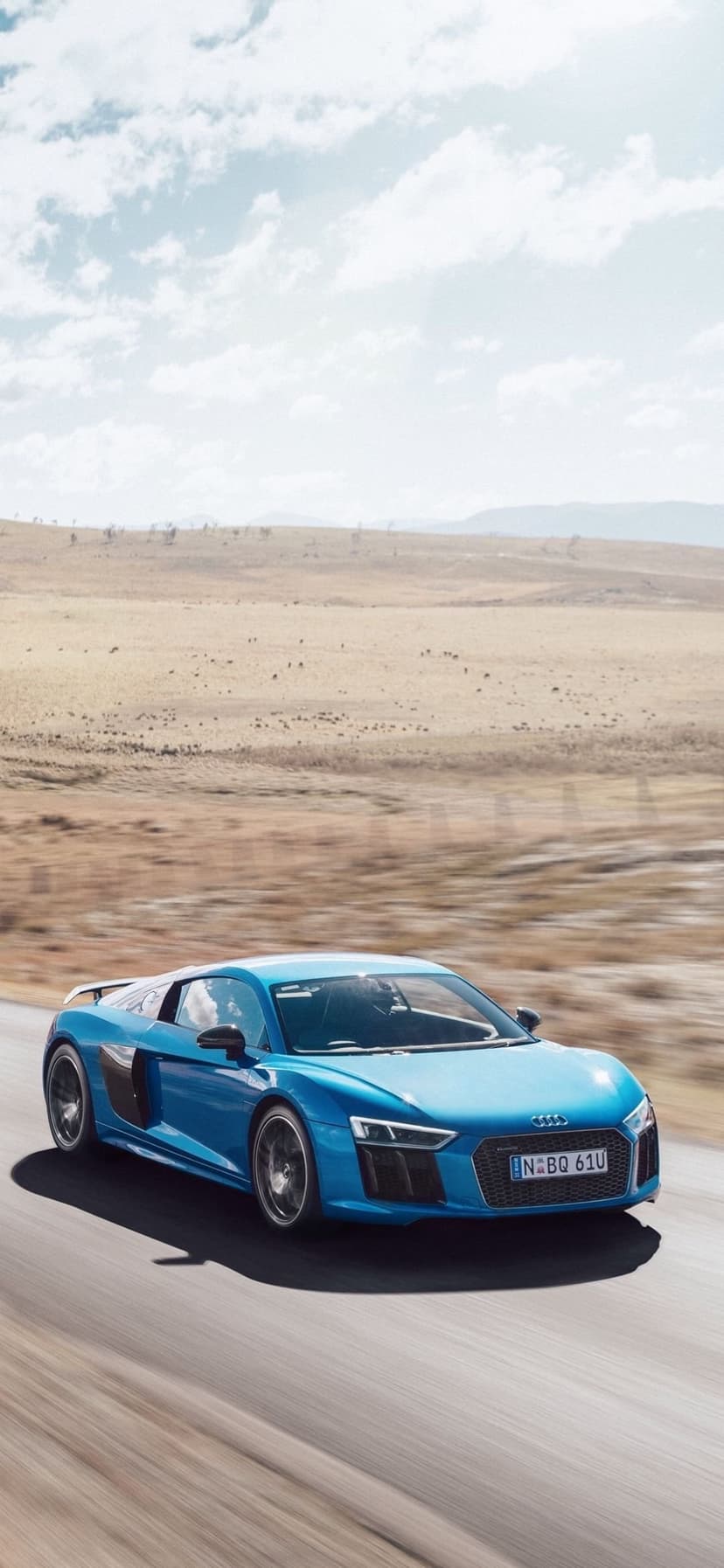 Blue Audi R8 sports car driving on a desert highway through dry plains and hills under a cloudy summer sky.