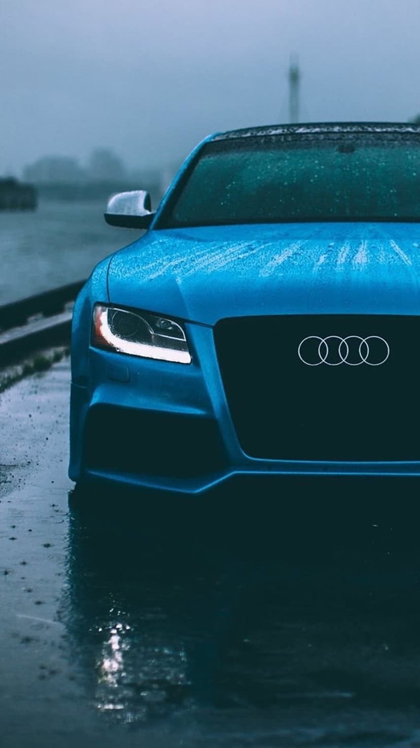 Blue Audi grille with silver rings sits on a rain-slicked road with glowing headlights and water beads.