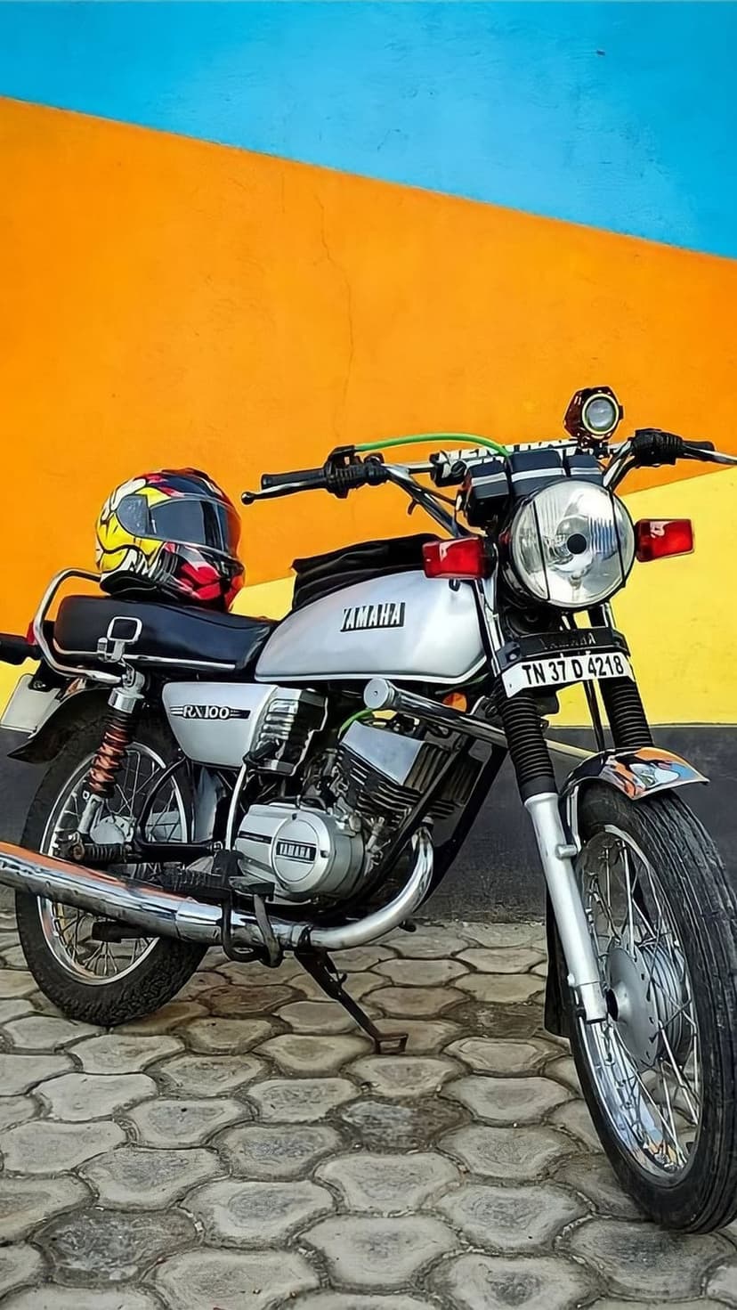 Yamaha RX100 motorcycle with a helmet on its seat parked on a stone pavement against a blue and orange wall.