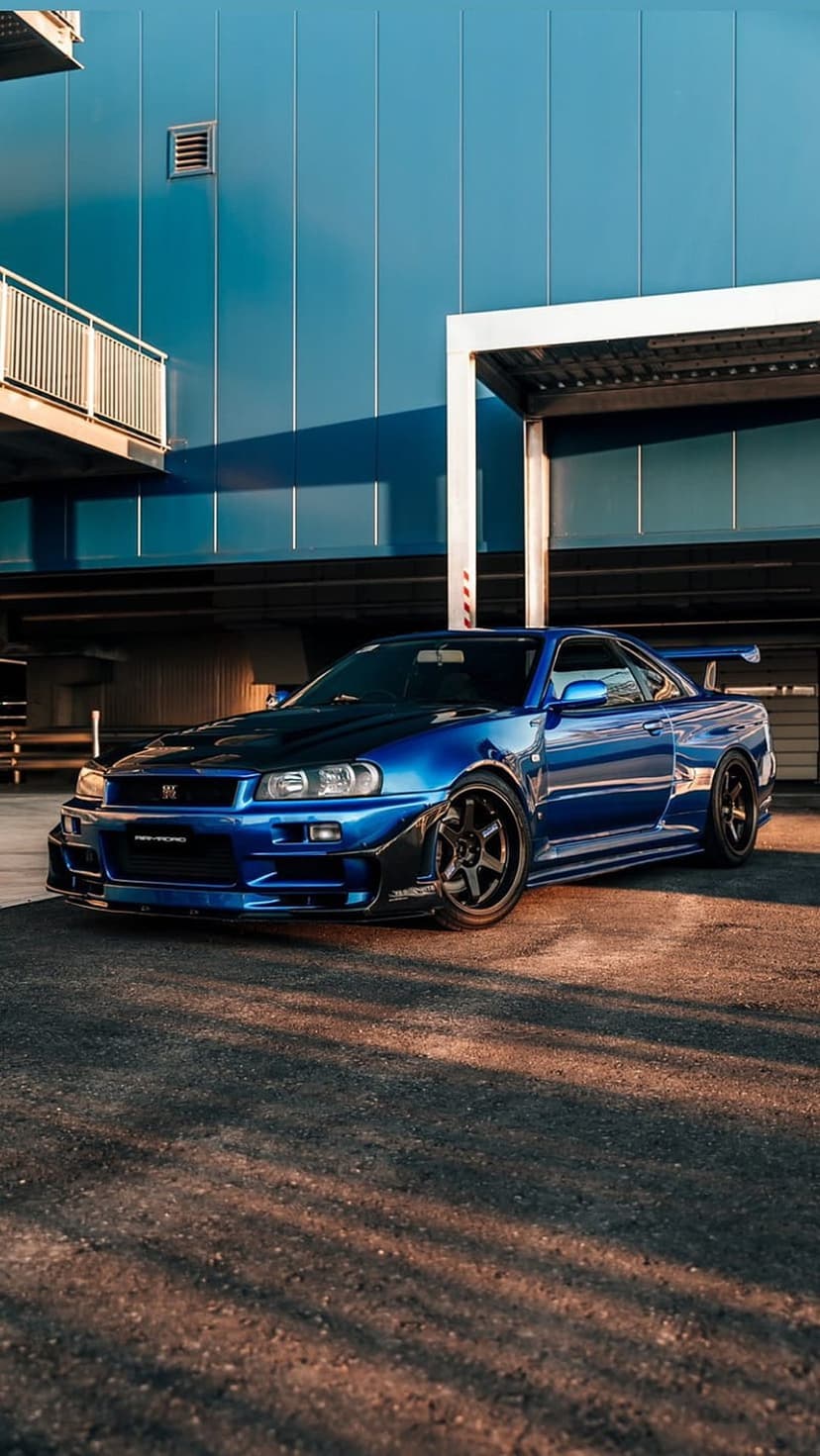 Blue Nissan GT-R R34 with a widebody kit and rear wing parked on a warm-toned street by blue urban buildings.