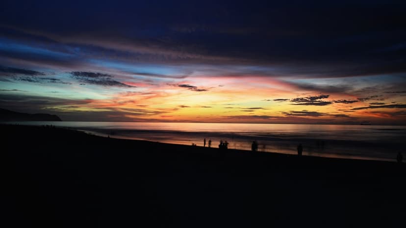 Tiny silhouetted figures walk along a dark sandy beach under a vibrant orange and purple twilight sky with waves.