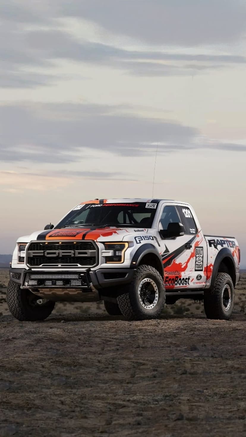 Ford F-150 Raptor race truck with custom graphics parked on desert sand under a cloudy sky during a Baja event.