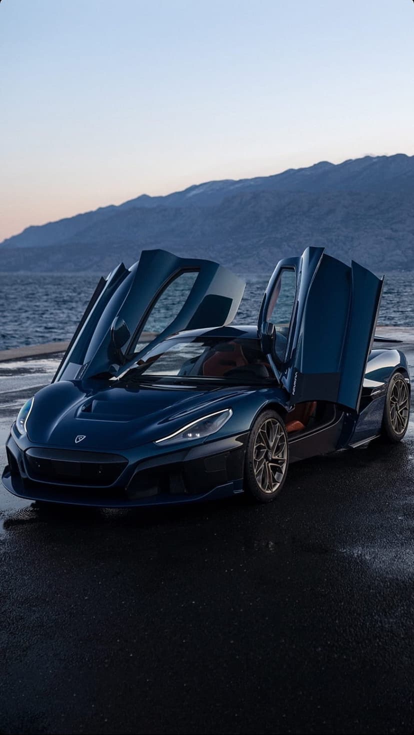 Blue Rimac Nevera hypercar with open butterfly doors parked near a tranquil sea under a clear mountain horizon.