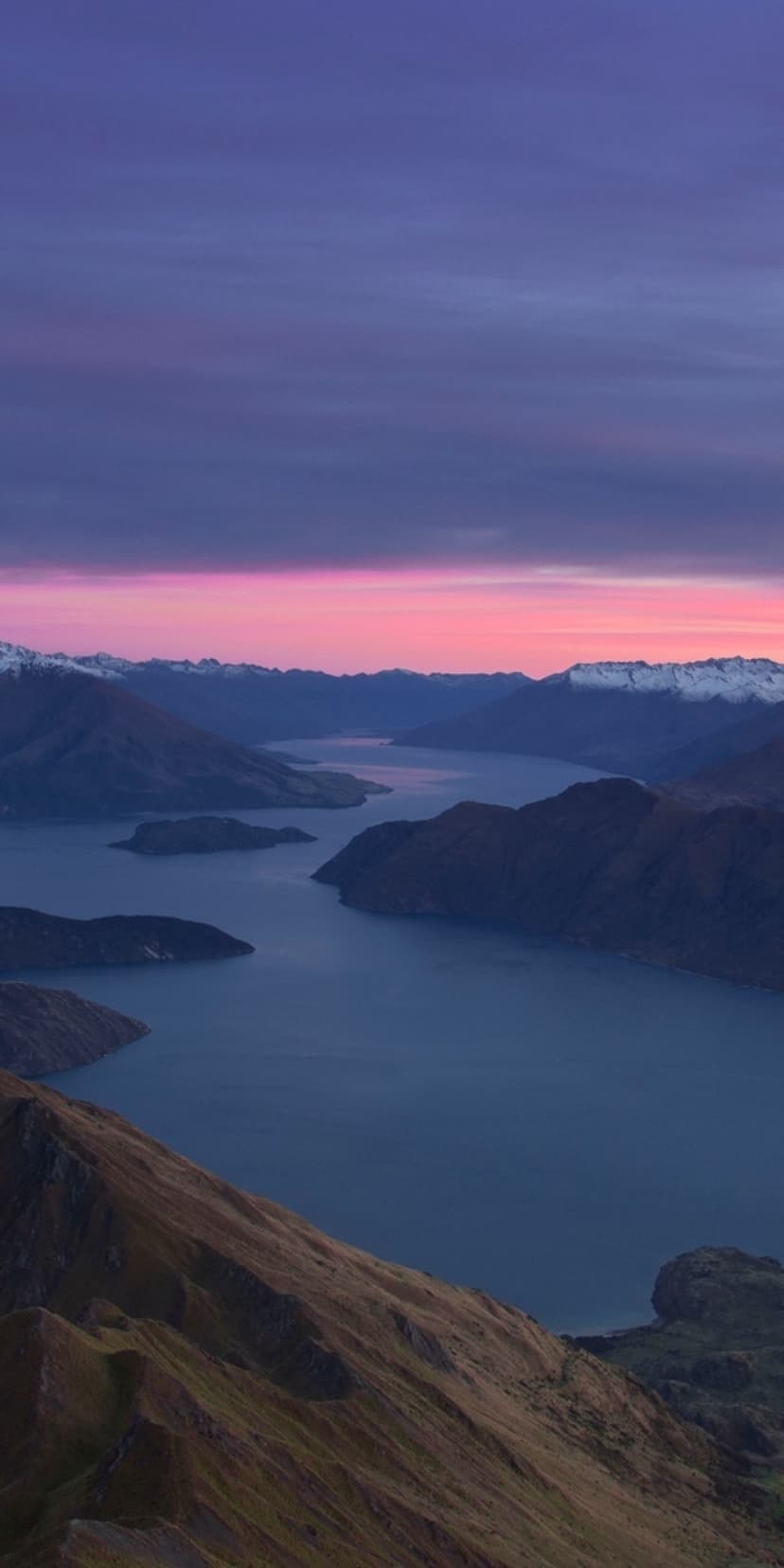 Rugged mountain ridges and a winding lake reflect a vibrant pink and purple twilight sky with snowy peaks.