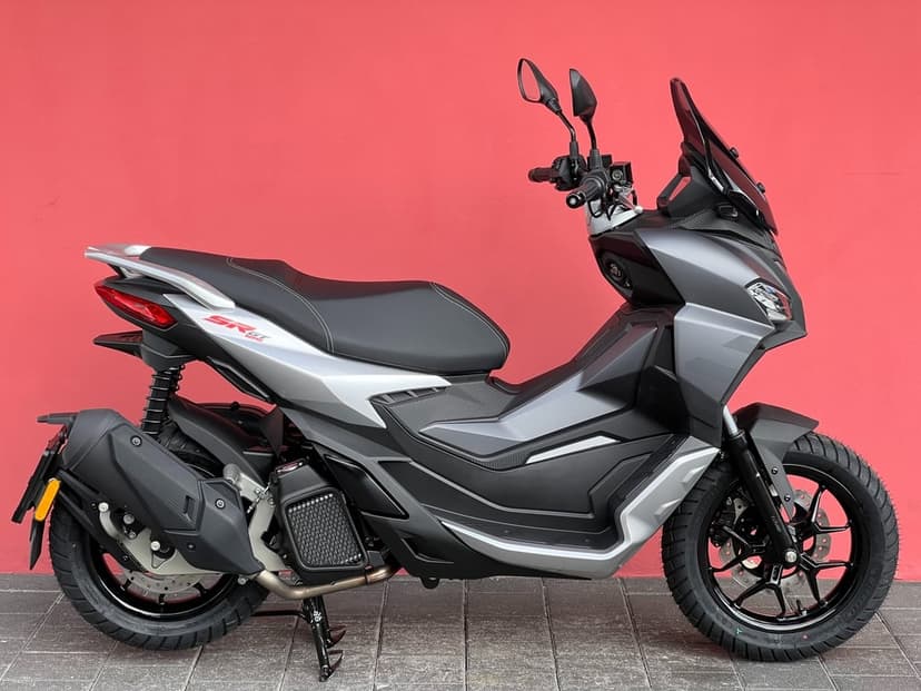 Grey and silver Aprilia SR GT 125 scooter parked sideways against a solid vibrant red wall background.