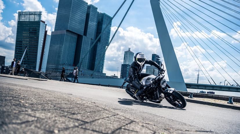 A biker in black leather leans into a turn on a grey city street with blue skyscrapers and a white bridge behind.