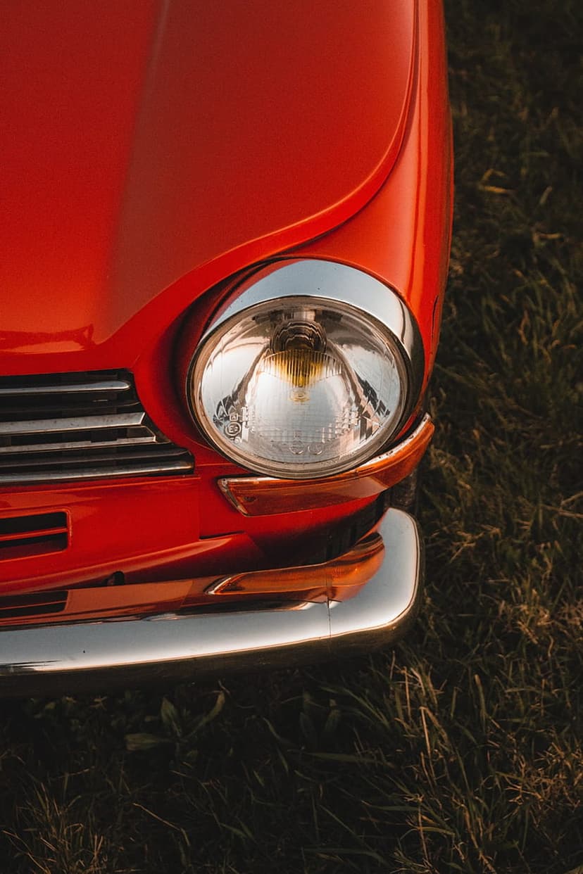 Red vintage car headlight and chrome bumper reflect sunlight while parked in a textured green grassy field.