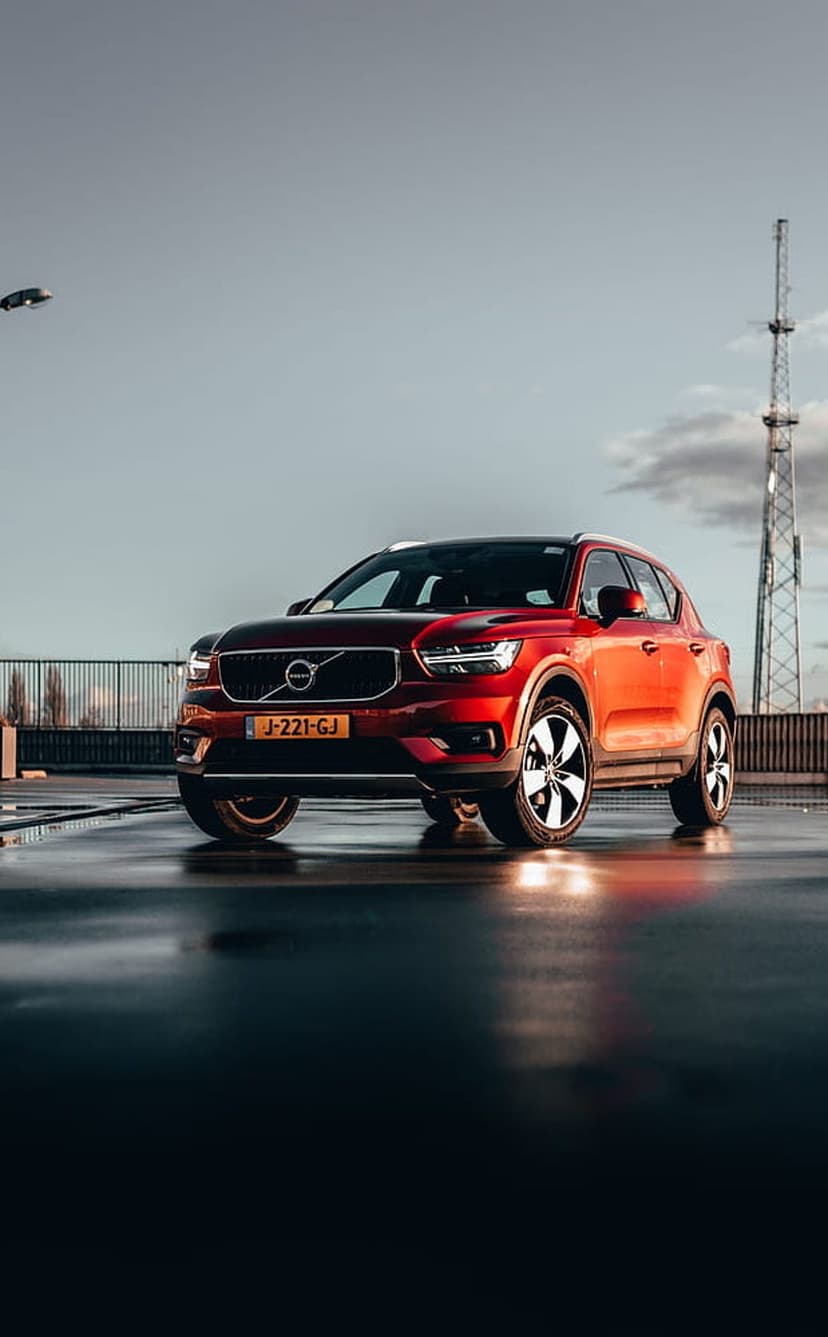 Red Volvo XC40 SUV reflects on a damp urban road against a bright overcast sky with a tall communication mast.