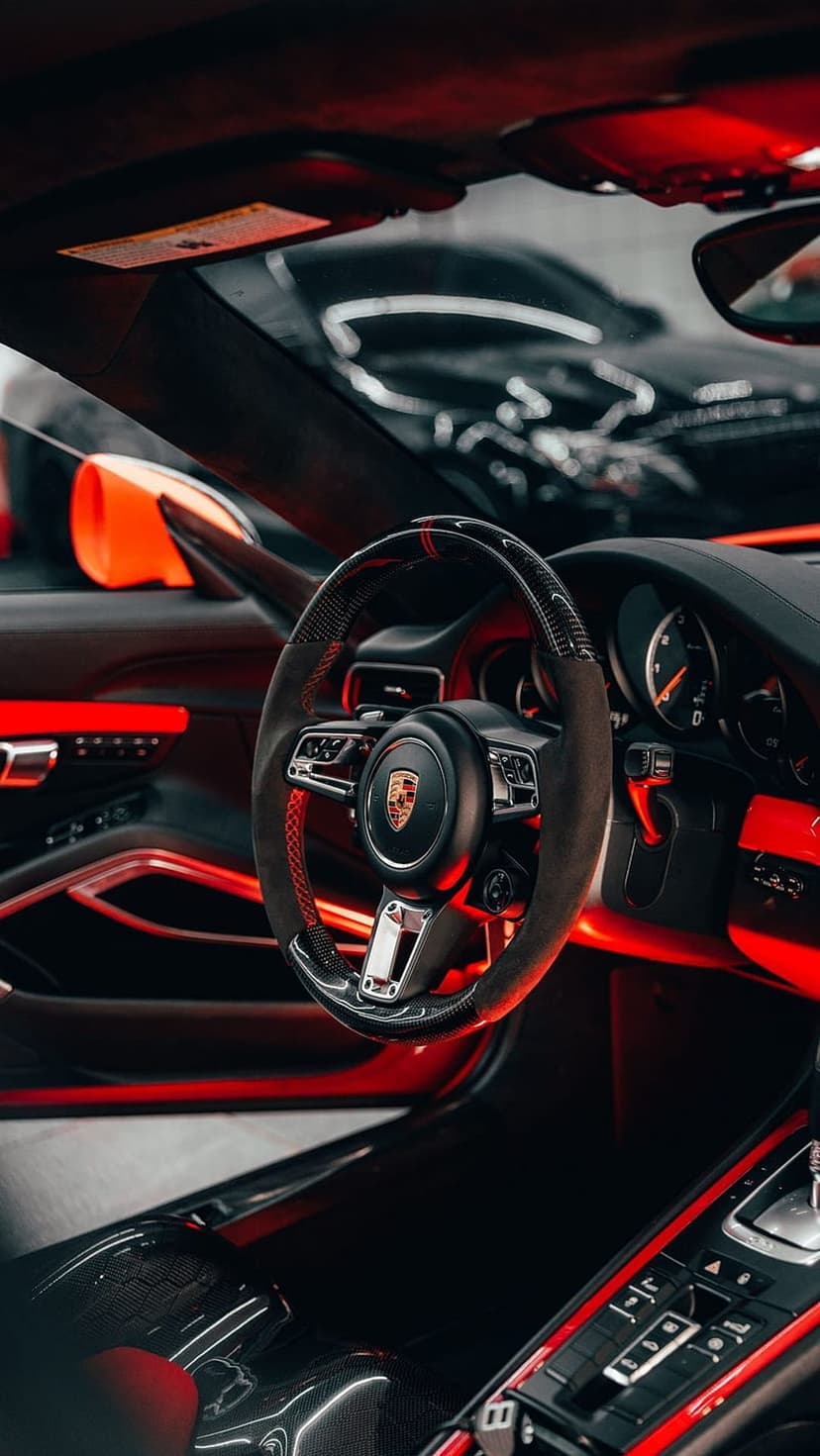 Porsche steering wheel with red stitching and emblem illuminated by scarlet ambient lights in a dark car cabin.