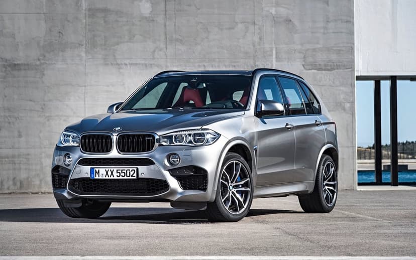 Silver BMW X5 M performance SUV parked in front of a concrete wall with red interior and blue calipers.