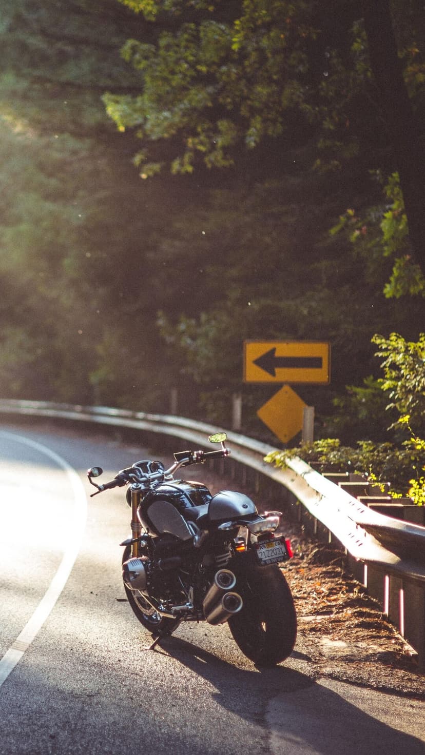 Motorcycle parked on a curving road at sunset with green trees and a yellow road sign in the background.