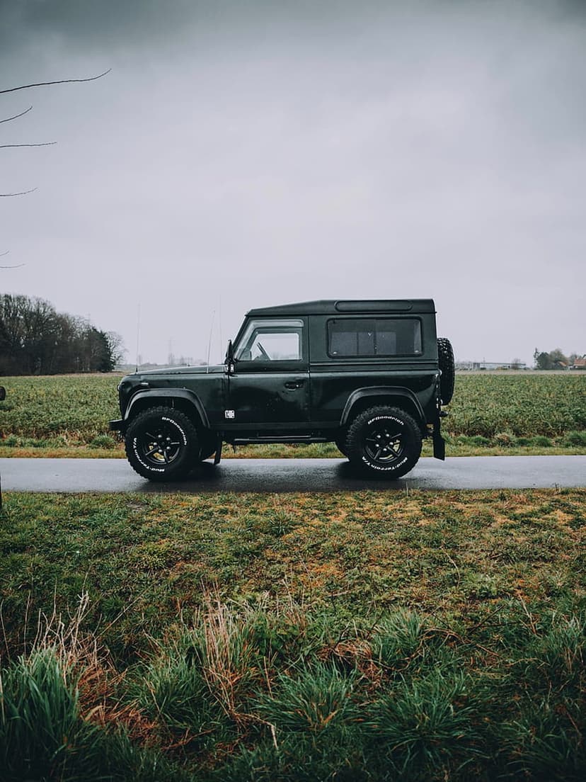 Dark green Land Rover Defender 4x4 parked on a glistening wet asphalt road surrounded by lush green grassy fields.