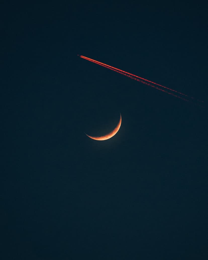 Slender orange crescent moon glows beneath a vivid red jet contrail streaking across a dark black night sky.
