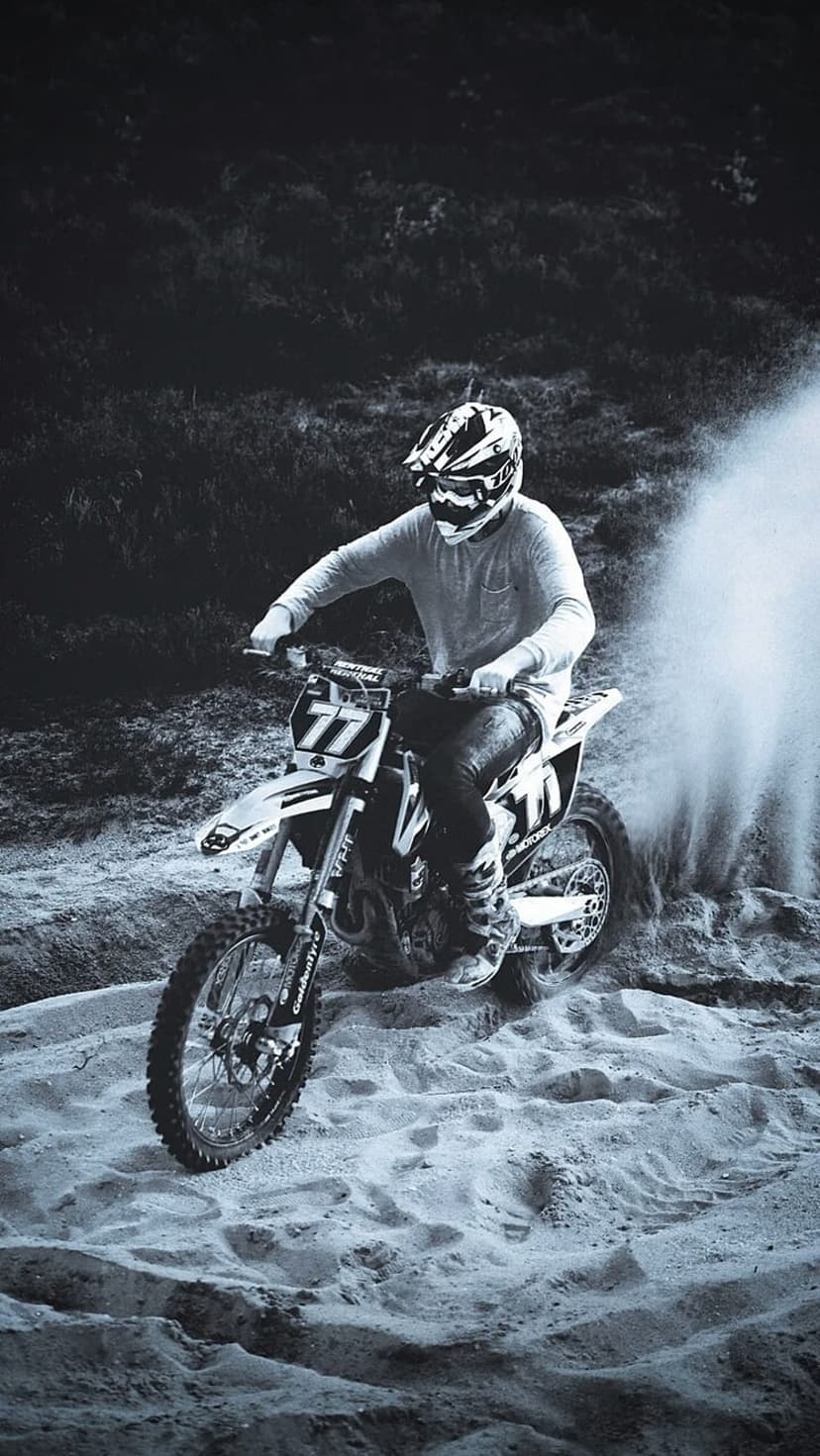 Motocross rider in gear leans into a turn as their motorcycle kicks up a large spray of sand and grit.