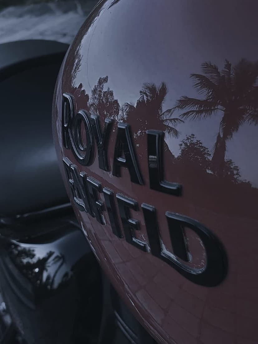 Royal Enfield red fuel tank with embossed silver logo reflecting palm trees under a sunset sky during twilight.
