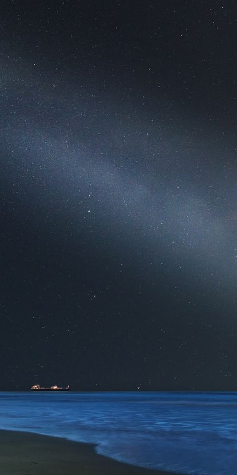 Small illuminated boat floating on a calm dark ocean under a starry sky and a visible Milky Way galaxy.