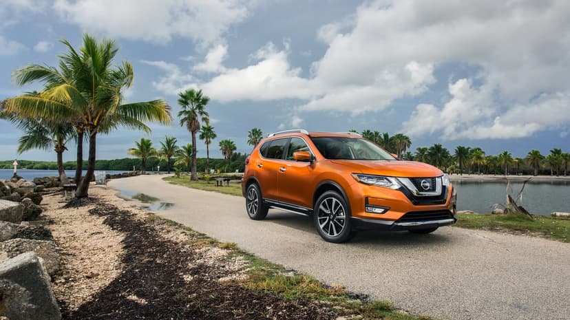 Orange Nissan Rogue SUV parked on a paved tropical path lined with palm trees under a bright cloudy sky.