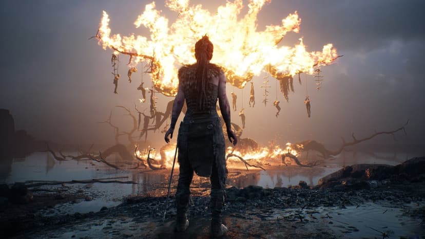 Woman in tribal attire stands before a large tree engulfed in bright orange flames under a dark misty sky.