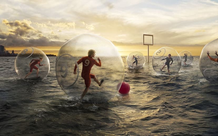 Runners inside transparent spheres compete on the ocean surface under a bright orange sunset with a distant city.