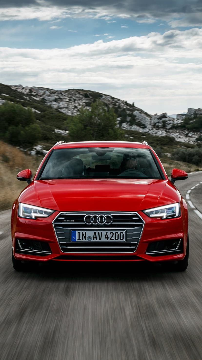 Red Audi A4 Avant wagon with glowing LED headlights driving on a winding asphalt road through rocky hills.