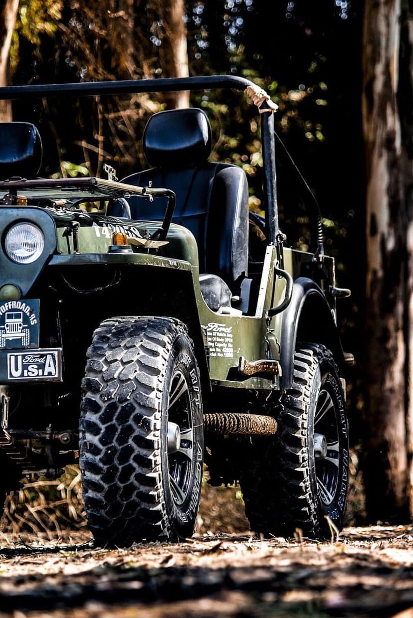 Vintage olive green Jeep with off-road tires parked on a dirt path under dappled sunlight in a lush forest.