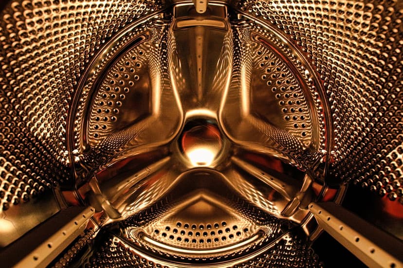 Perforated stainless steel washing machine drum interior with golden lighting and a central agitator structure.