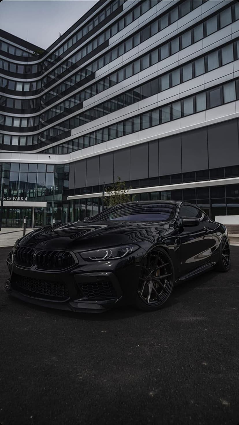 Black BMW M8 coupe parked on asphalt in front of a modern office building with white horizontal accents.