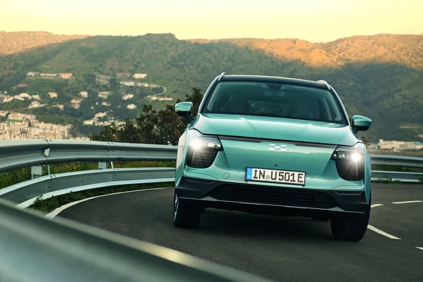 Teal electric SUV with headlights on driving on a curved mountain road with hills in the background.